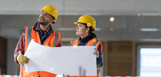 Two engineers holding blueprints, inside a building still under construction.